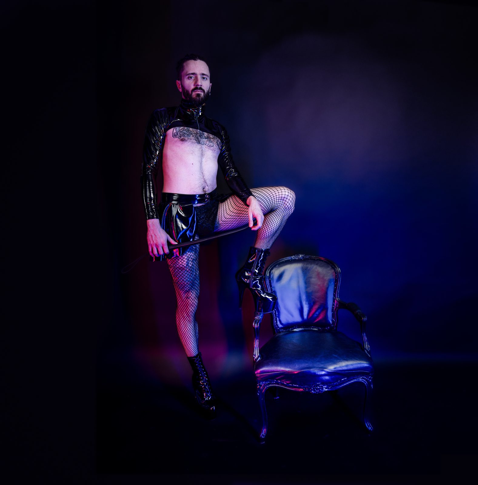 Princess Boy standing beside a chair in latex and fishnets, holding a riding crop