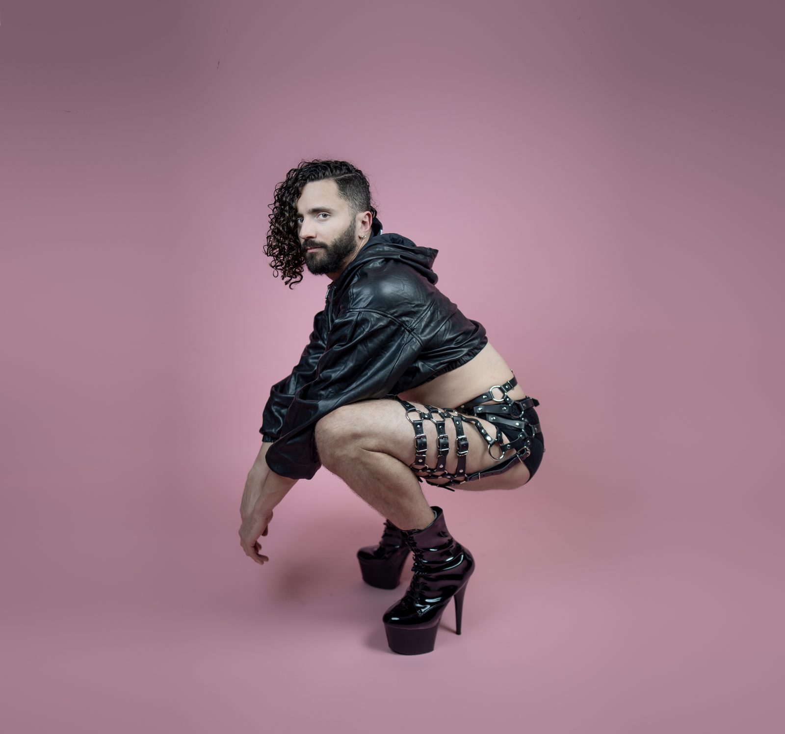 Princess Boy crouched in heels, a cropped leather jacket and a leg harness, against a pink background